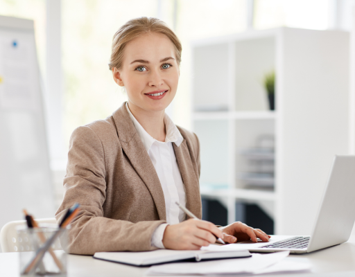 Secretária de governança sorrindo em ambiente corporativo, escrevendo em caderno ao lado de um notebook