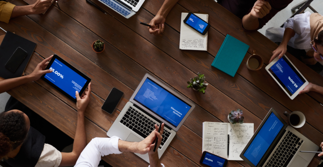 Pessoas reunidas em torno de uma mesa com notebooks e tablets, em uma reunião digital com uso de tecnologia.